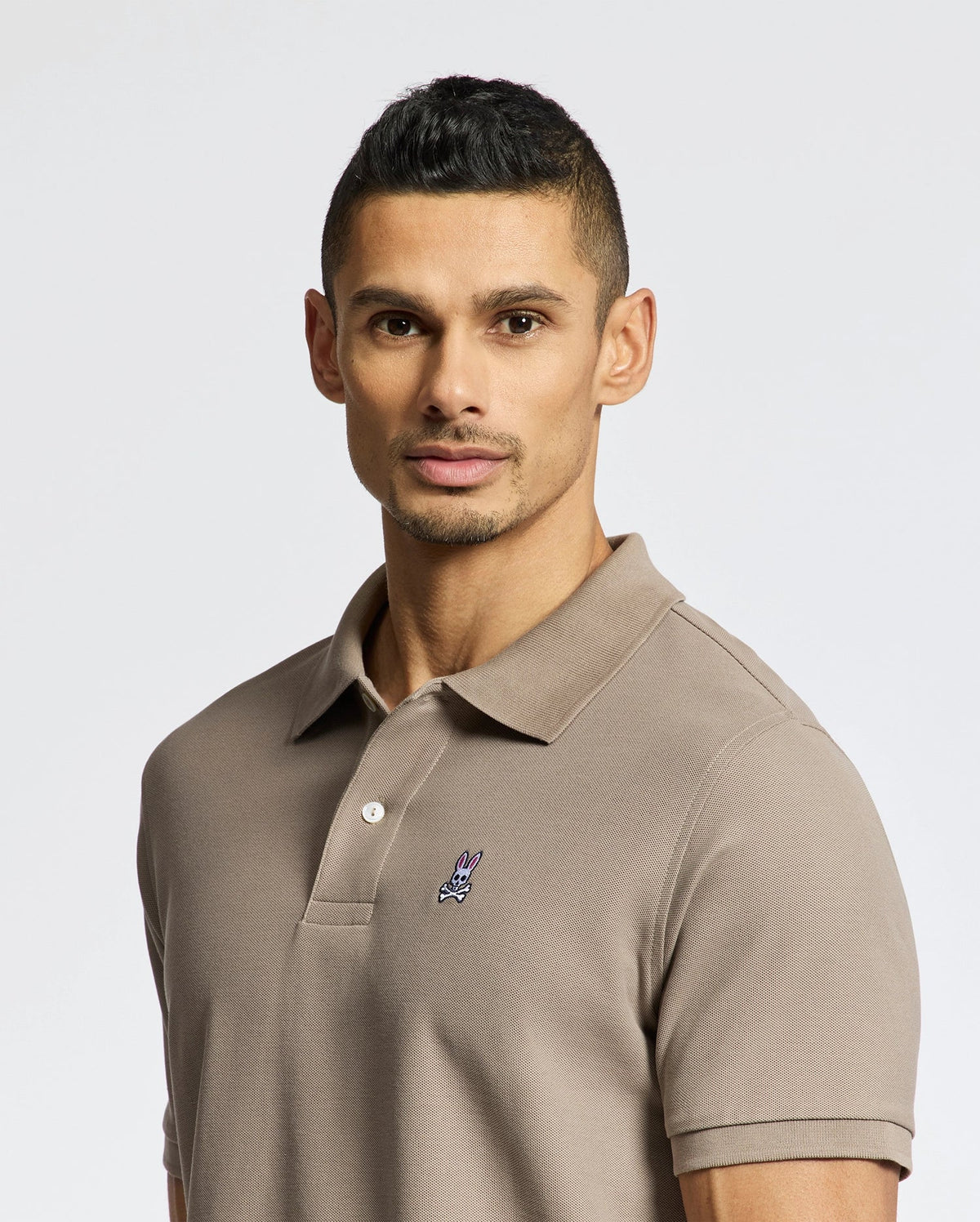 A man with short dark hair wears the soft taupe Mens Classic Pique Polo Shirt - B6K001CRPC by Psycho Bunny, featuring a small graphic on the chest. He looks directly at the camera against a plain white background.