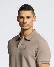 A man with short dark hair wears the soft taupe Mens Classic Pique Polo Shirt - B6K001CRPC by Psycho Bunny, featuring a small graphic on the chest. He looks directly at the camera against a plain white background.
