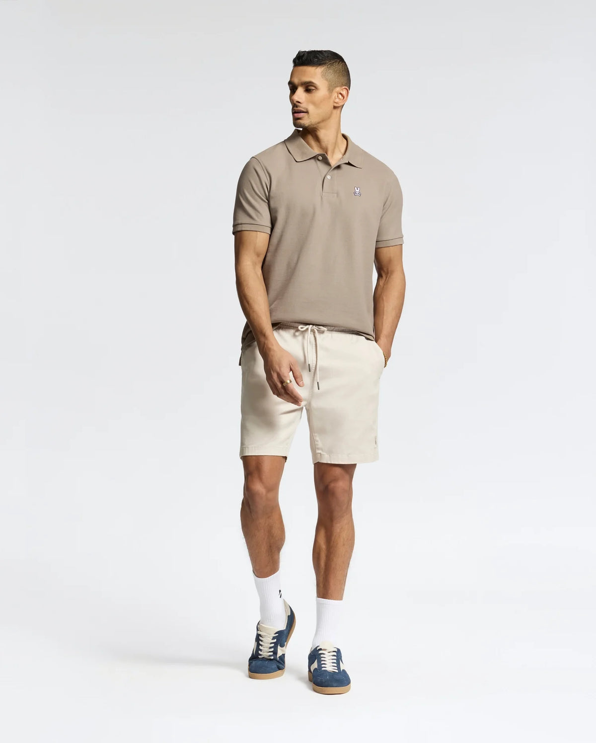 A man in a light brown Psycho Bunny Men's Classic Pique Polo Shirt - B6K001CRPC with beige shorts, white socks, and dark sneakers stands with one foot forward and hands in pockets against a plain white background.