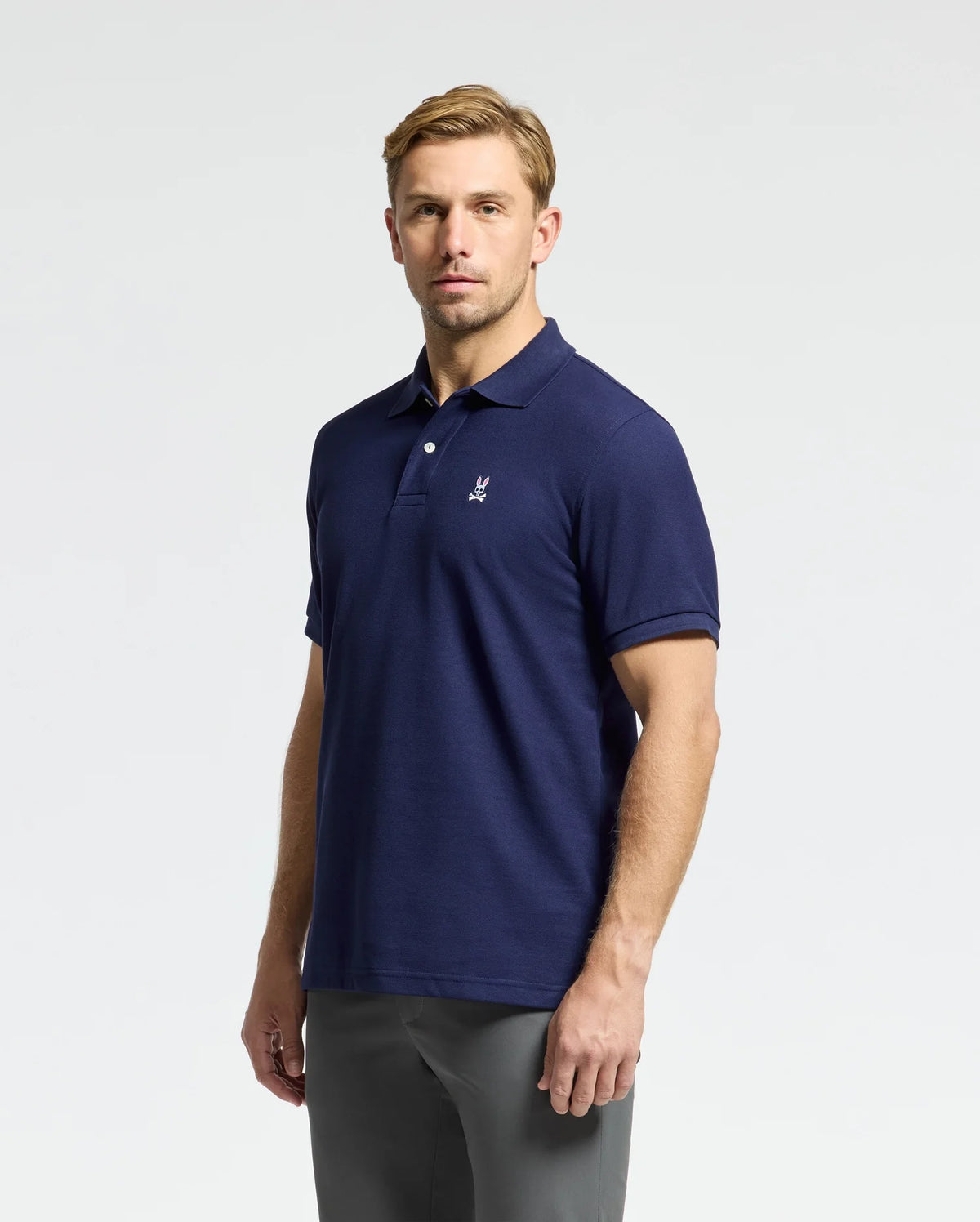 A man models the Psycho Bunny MENS CLASSIC PIQUE POLO SHIRT - B6K001CRPC, featuring a deep blue color and a subtle logo on the chest, against a simple white background. Made from high-quality pima cotton, he gazes directly at the camera with an expressionless face.