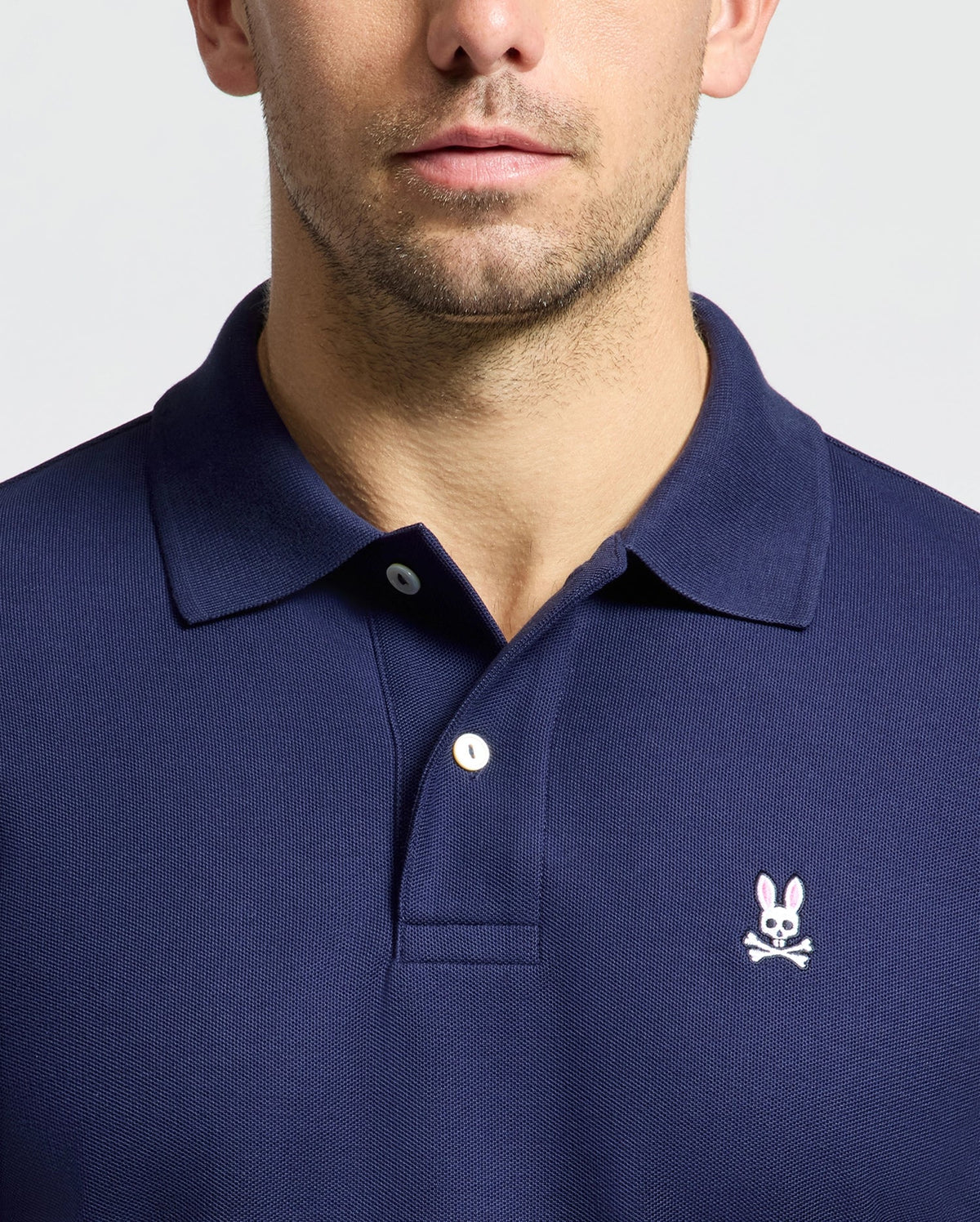 A person wearing a Psycho Bunny MENS CLASSIC PIQUE POLO SHIRT - B6K001CRPC in navy blue, crafted from soft Pima cotton, features a small embroidered logo of a white rabbit with crossed bones beneath it, positioned over the left side of the chest. The image focuses on the upper torso and part of the face.