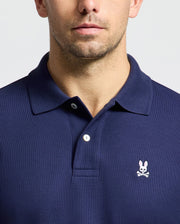 A person wearing a Psycho Bunny MENS CLASSIC PIQUE POLO SHIRT - B6K001CRPC in navy blue, crafted from soft Pima cotton, features a small embroidered logo of a white rabbit with crossed bones beneath it, positioned over the left side of the chest. The image focuses on the upper torso and part of the face.