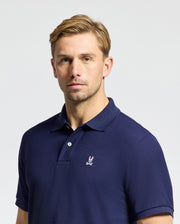 A man with short blond hair is wearing the MENS CLASSIC PIQUE POLO SHIRT - B6K001CRPC by Psycho Bunny, crafted from soft pima cotton and featuring a small embroidered rabbit logo. He stands against a plain background, looking directly at the camera with a neutral expression.