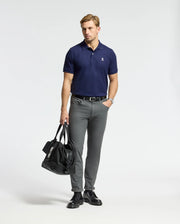 A man wearing a Psycho Bunny MENS CLASSIC PIQUE POLO SHIRT - B6K001CRPC in navy pairs it with gray pants and holds a black duffel bag. He accessorizes with a wristwatch while standing in front of a plain white background.