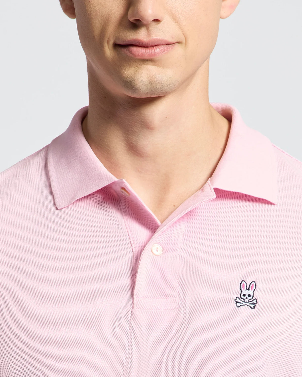 A person wears the Psycho Bunny MENS CLASSIC PIQUE POLO SHIRT - B6K001CRPC in light pink, featuring a collared, button design. Made from soft Pima cotton, it showcases an embroidered rabbit above crossbones logo on the chest against a plain light gray background.