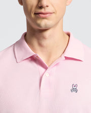 A person wears the Psycho Bunny MENS CLASSIC PIQUE POLO SHIRT - B6K001CRPC in light pink, featuring a collared, button design. Made from soft Pima cotton, it showcases an embroidered rabbit above crossbones logo on the chest against a plain light gray background.
