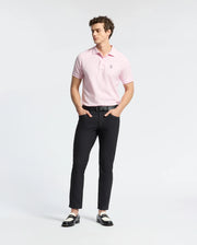 A person stands wearing a light pink Psycho Bunny MENS CLASSIC PIQUE POLO SHIRT - B6K001CRPC and black pants, hands in pockets. Two-tone loafers complete the look against a white background, highlighting the Pima cotton clothing.