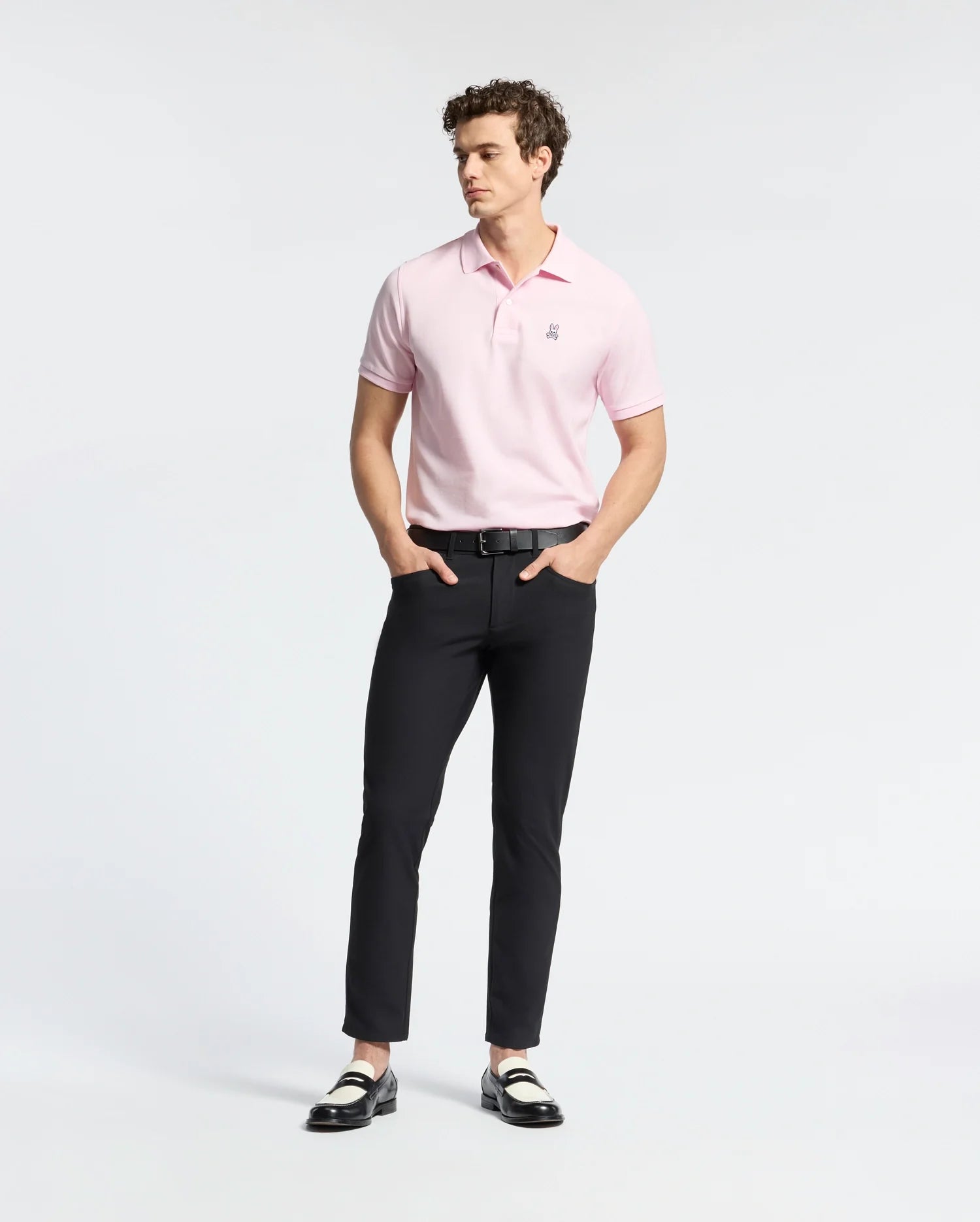 A person stands wearing a light pink Psycho Bunny MENS CLASSIC PIQUE POLO SHIRT - B6K001CRPC and black pants, hands in pockets. Two-tone loafers complete the look against a white background, highlighting the Pima cotton clothing.