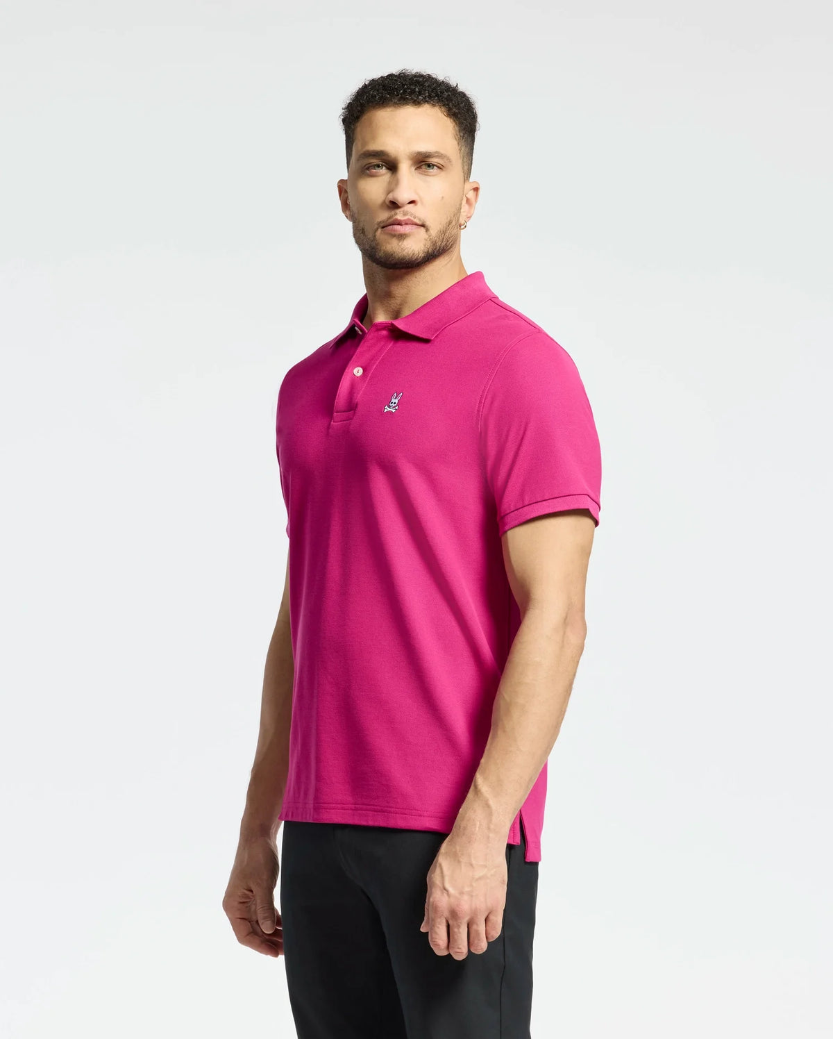 A man confidently stands against a plain white background wearing Psycho Bunny's vibrant pink B6K001CRPC classic piqué polo, featuring the iconic logo on his chest, paired with black pants.