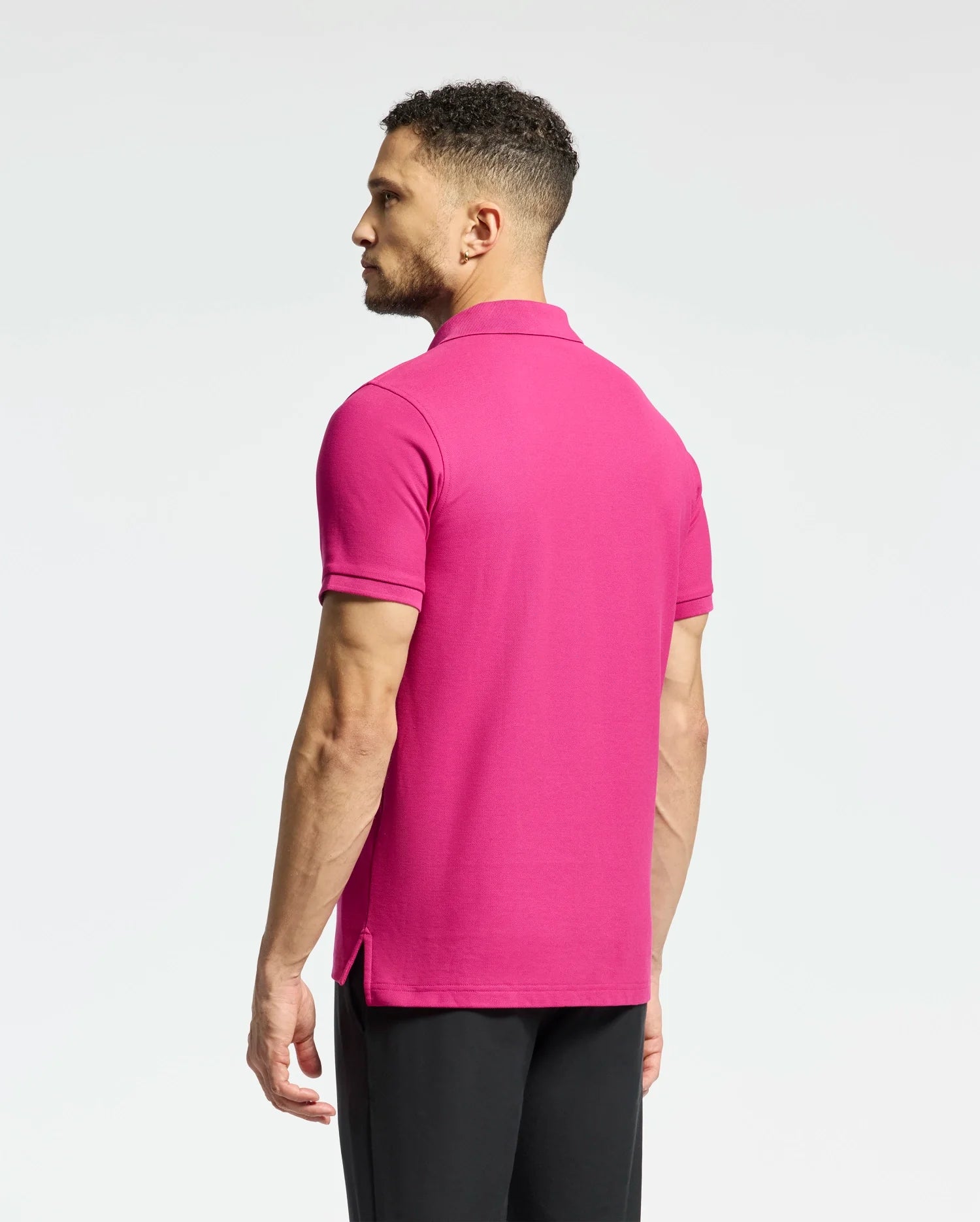 A person with short curly hair, facing away against a white background, wears black pants and the Psycho Bunny Men's Classic Piqué Polo Shirt in bright pink.