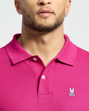 Sporting the Psycho Bunny Mens Classic Pique Polo Shirt (B6K001CRPC) in bright pink, featuring the iconic cartoon bunny with crossbones logo on the chest, a person is styled with a beard and small hoop earring against a light gray backdrop.
