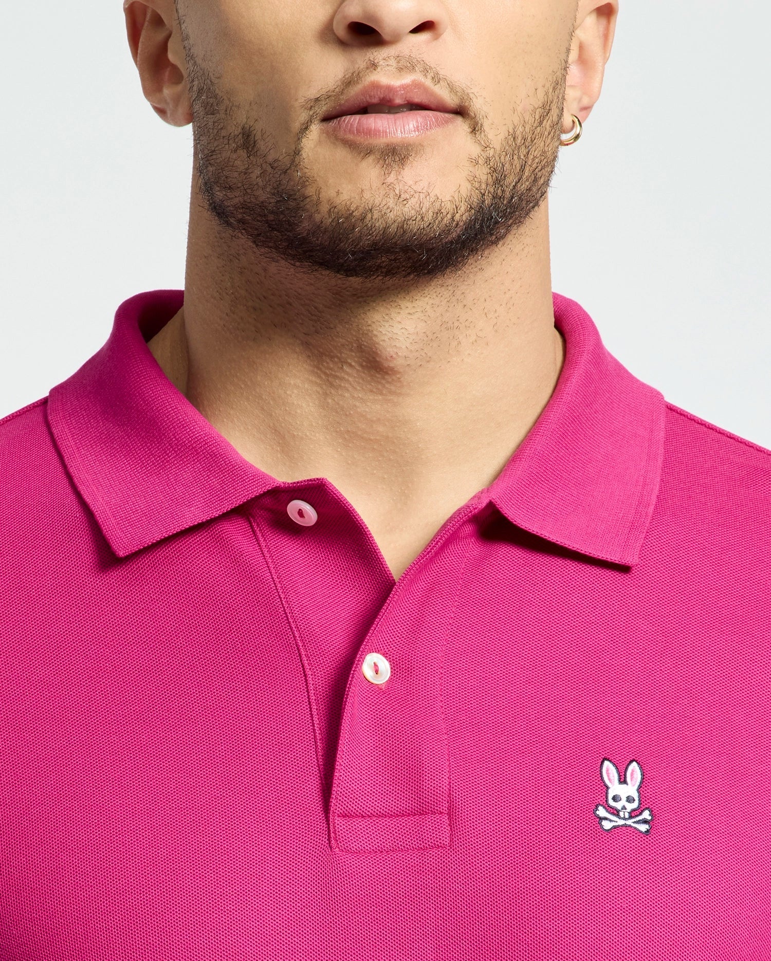 Sporting the Psycho Bunny Mens Classic Pique Polo Shirt (B6K001CRPC) in bright pink, featuring the iconic cartoon bunny with crossbones logo on the chest, a person is styled with a beard and small hoop earring against a light gray backdrop.