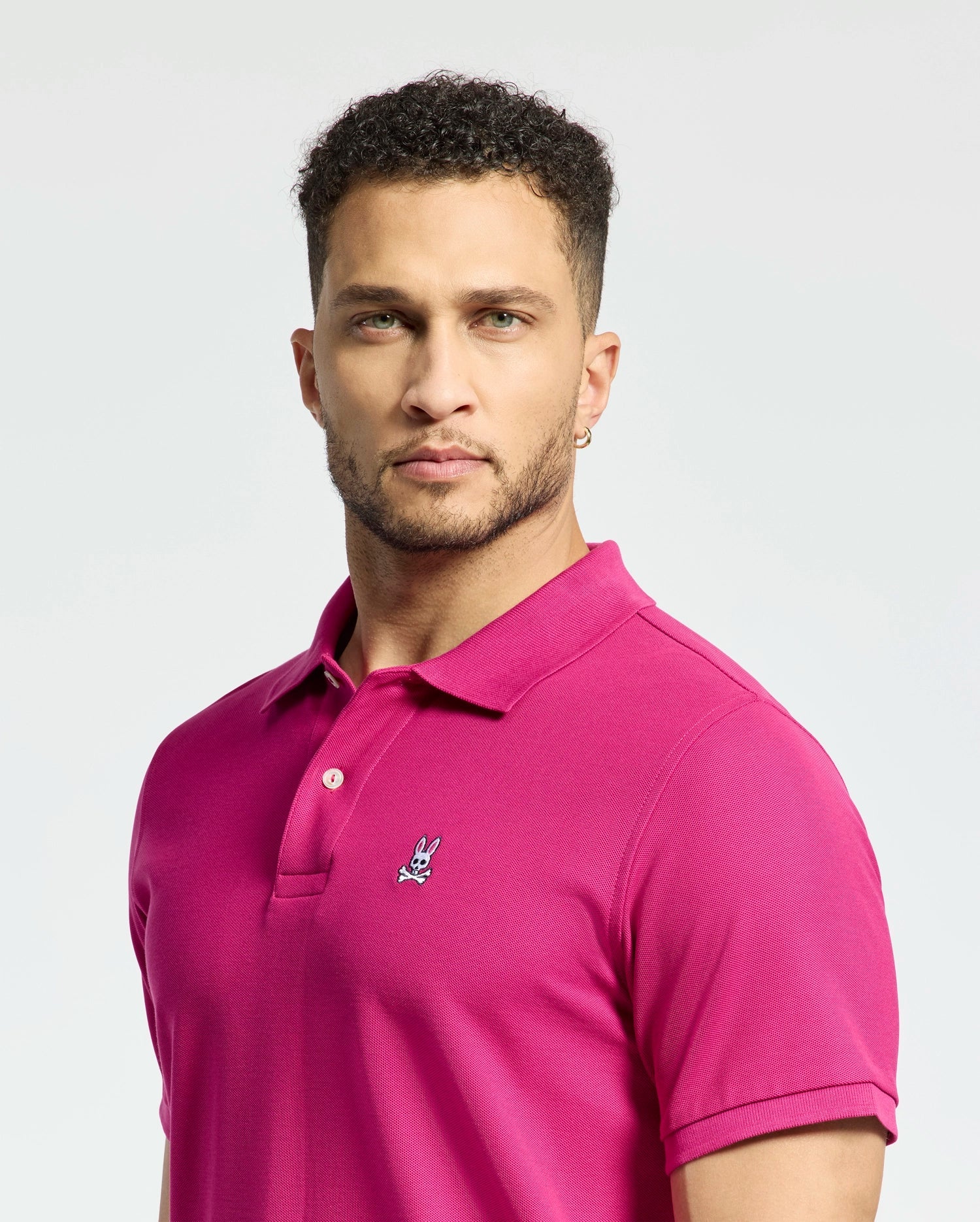 A man with short curly hair and a beard is wearing a bright pink Psycho Bunny Men's Classic Pique Polo Shirt (B6K001CRPC), made from Pima cotton, featuring the iconic embroidered rabbit skull and crossbones logo on the chest, set against a plain light background.