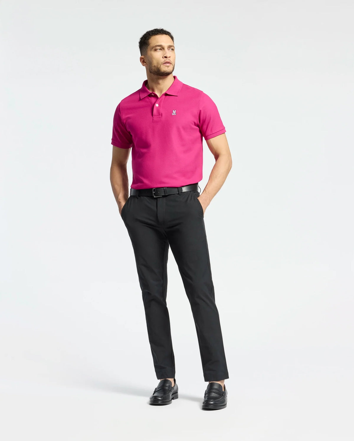 A person stands with their hands in pockets, wearing a bright pink Psycho Bunny MENS CLASSIC PIQUE POLO SHIRT (B6K001CRPC), black trousers, and shoes against a white background.