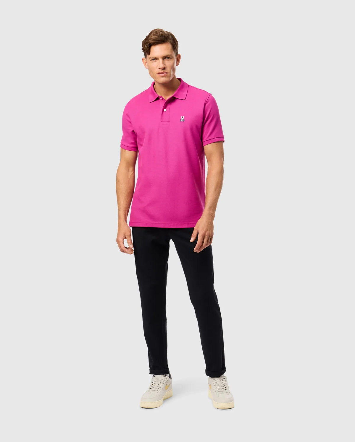 A person wearing the Psycho Bunny MENS CLASSIC PIQUE POLO SHIRT - B6K001D200 in a vibrant pink hue stands against a plain background. Paired with black pants and white sneakers, the 100% Pima cotton fabric exudes subtle elegance as they gaze ahead with a neutral expression.