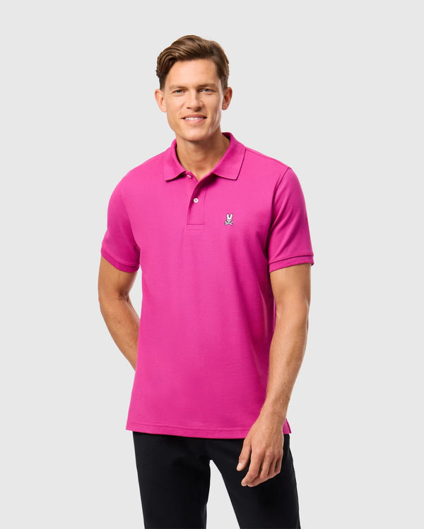 Wearing the Psycho Bunny MENS CLASSIC PIQUE POLO SHIRT in a vibrant pink hue, made from 100% Pima cotton and adorned with a small embroidered logo on the left chest, a person stands against a plain light gray background in black pants, smiling softly. Made in Peru.