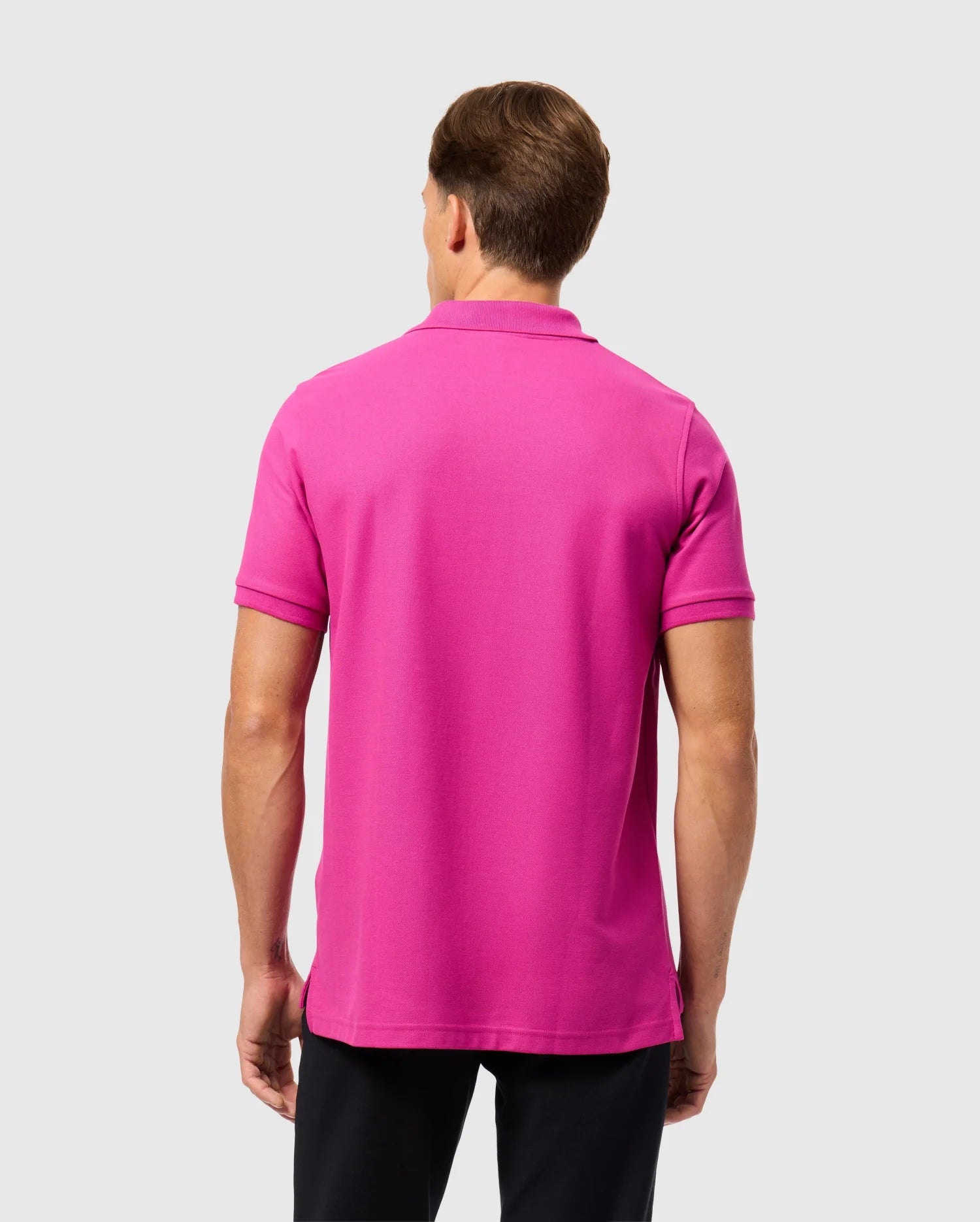 A man, facing away from the camera, is showcasing a vibrant pink Psycho Bunny Men's Classic Pique Polo Shirt - B6K001D200 made from 100% Pima cotton. He pairs it with black pants against a plain gray background, perfectly capturing understated elegance. Crafted in Peru, this garment effortlessly combines comfort and style in one sleek package.
