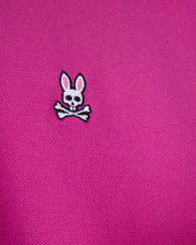 A close-up of the pink fabric from the Psycho Bunny Men's Classic Pique Polo Shirt - B6K001D200, crafted in Peru from 100% Pima cotton, showcasing a small emblem with a white skull and crossbones adorned with pink-eared bunny ears above.