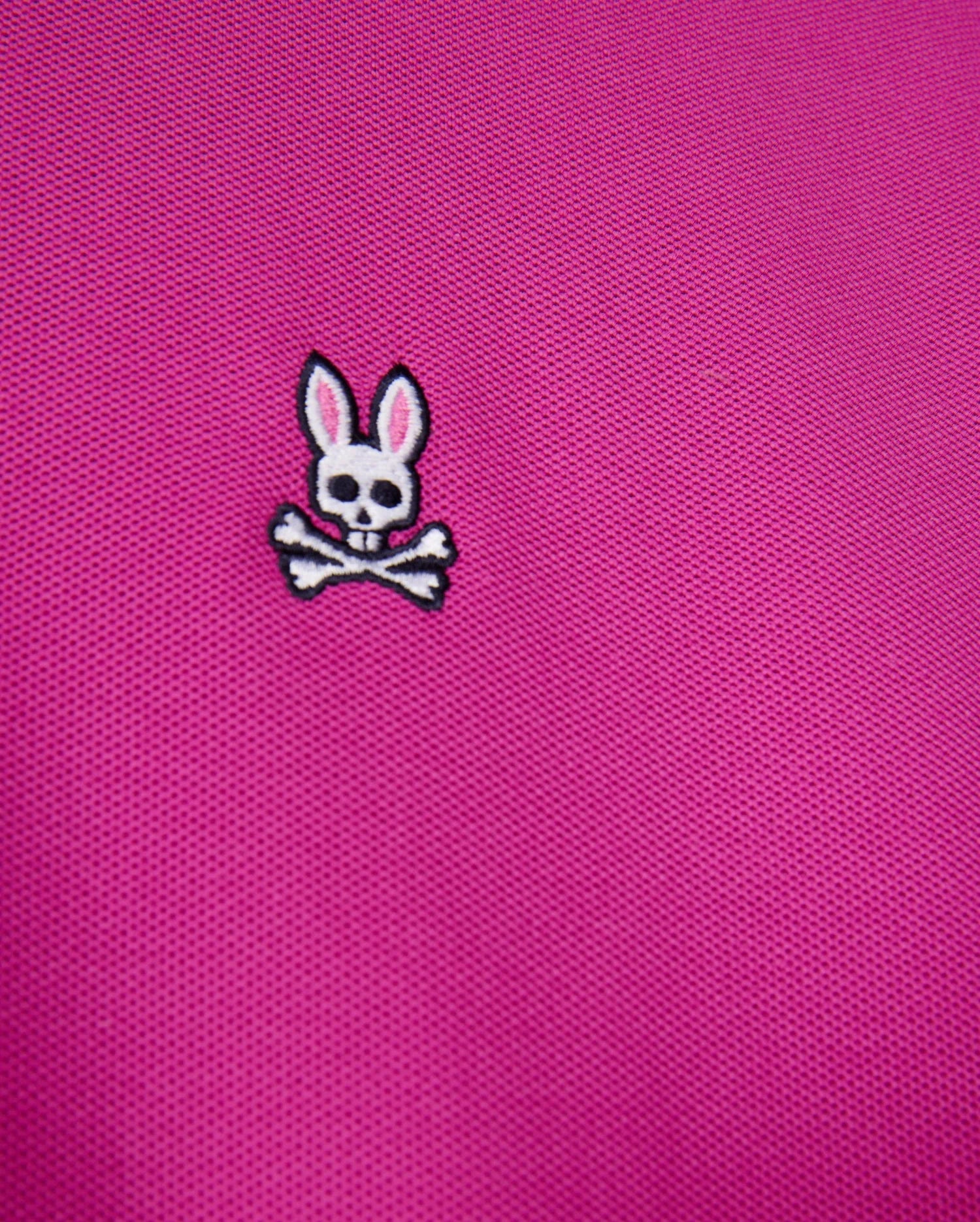 A close-up of the pink fabric from the Psycho Bunny Men's Classic Pique Polo Shirt - B6K001D200, crafted in Peru from 100% Pima cotton, showcasing a small emblem with a white skull and crossbones adorned with pink-eared bunny ears above.