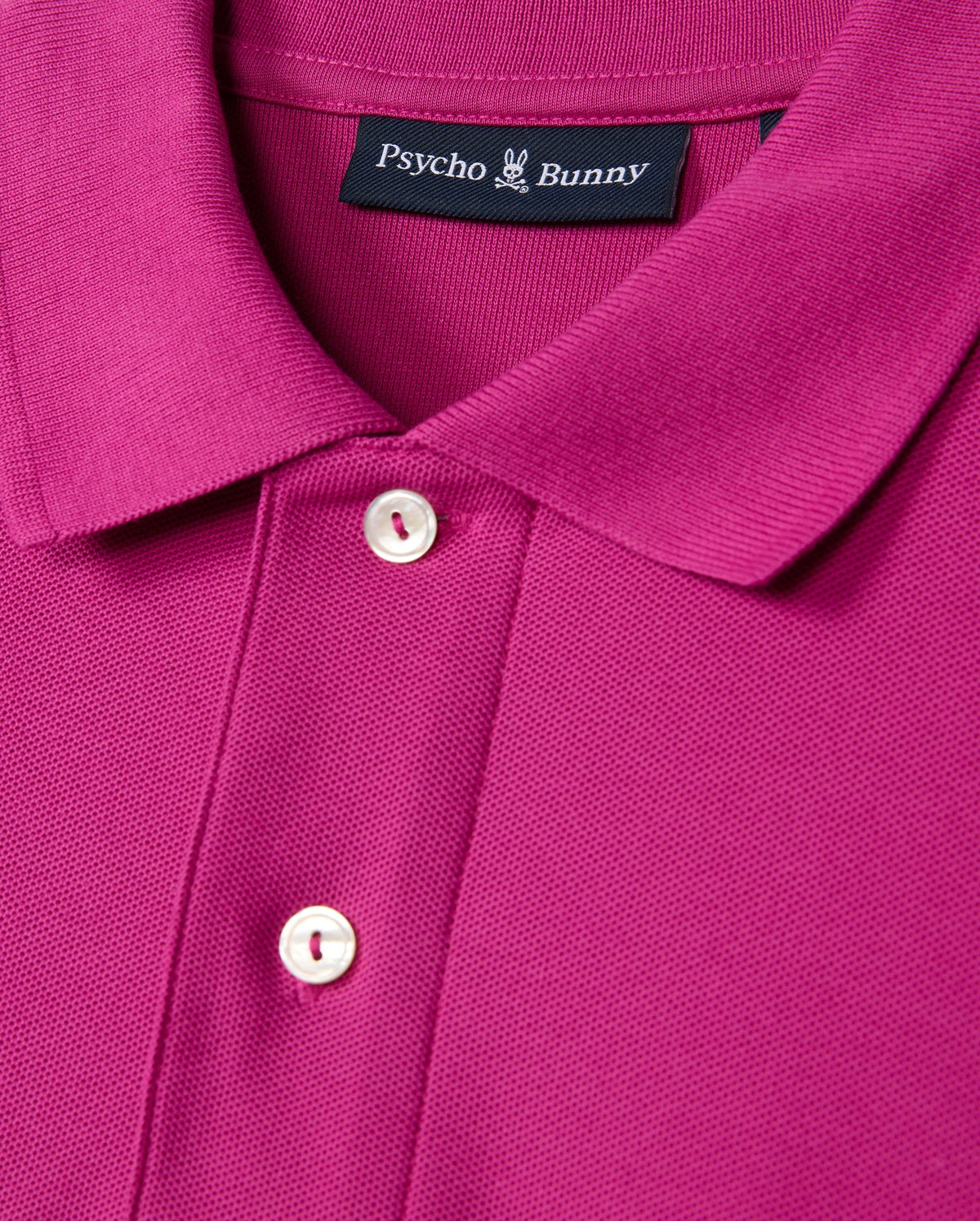 Close-up of a magenta "MENS CLASSIC PIQUE POLO SHIRT - B6K001D200" featuring a folded collar and two white buttons. Inside the collar, the label displays "Psycho Bunny" with their signature bunny logo above it. Crafted in Peru from 100% Pima cotton, this polo offers unparalleled comfort and style.