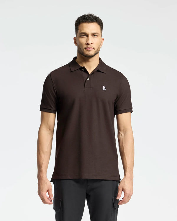 A man stands against a plain background wearing a Psycho Bunny MENS CLASSIC PIQUE POLO SHIRT - B6K001D200, featuring rich diamond-knit piqué fabric and a small chest emblem. His classic polo complements his dark pants as he gazes forward, short curly hair framing his neutral expression.