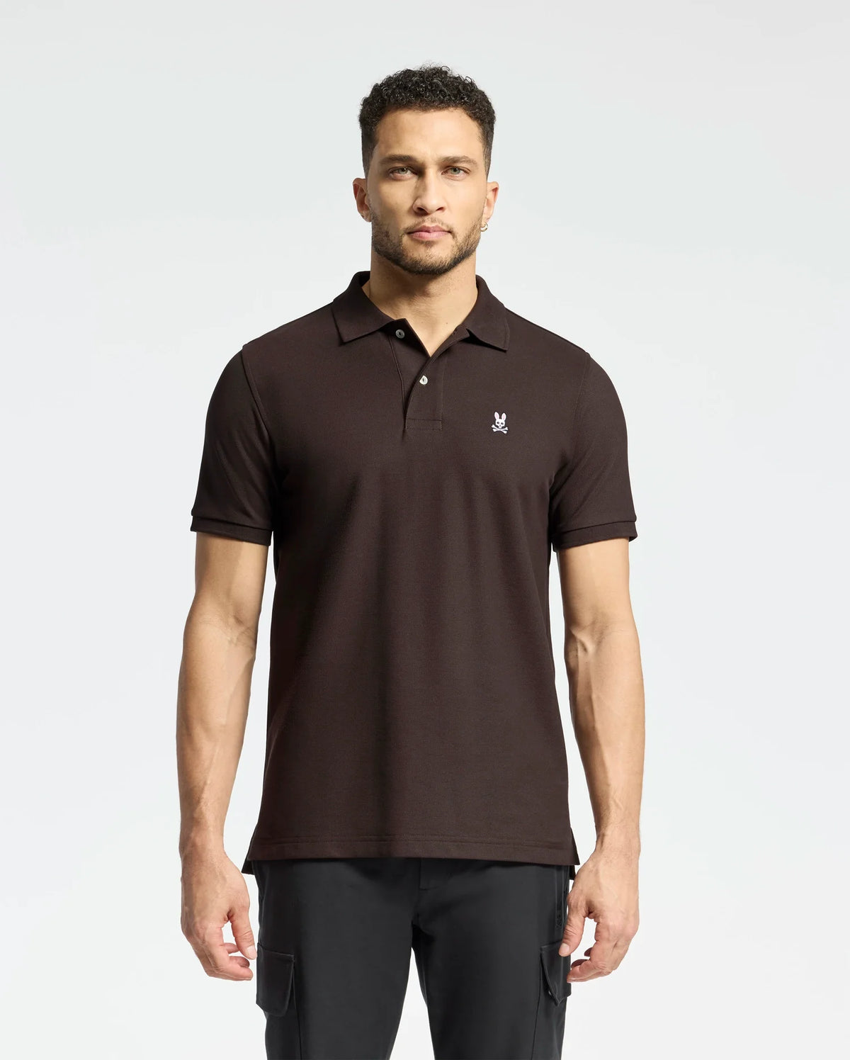 A man stands against a plain background wearing a Psycho Bunny MENS CLASSIC PIQUE POLO SHIRT - B6K001D200, featuring rich diamond-knit piqué fabric and a small chest emblem. His classic polo complements his dark pants as he gazes forward, short curly hair framing his neutral expression.