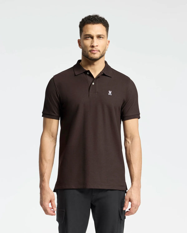 A man stands against a plain background wearing a Psycho Bunny MENS CLASSIC PIQUE POLO SHIRT - B6K001D200, featuring rich diamond-knit piqué fabric and a small chest emblem. His classic polo complements his dark pants as he gazes forward, short curly hair framing his neutral expression.