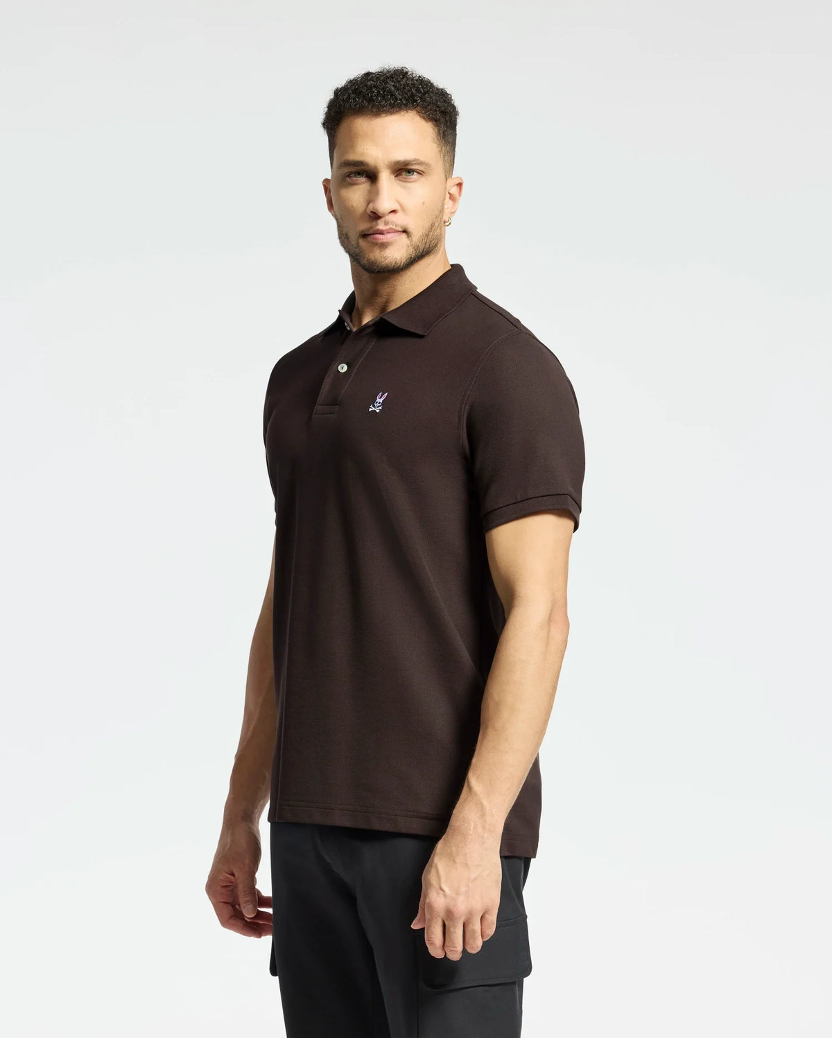A man stands against a plain white background wearing a Psycho Bunny MENS CLASSIC PIQUE POLO SHIRT - B6K001D200 in dark brown, made from luxurious Pima cotton with a small embroidered logo on the chest, paired with black pants.