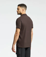 A man with short curly hair wears a Psycho Bunny MENS CLASSIC PIQUE POLO SHIRT - B6K001D200 and black pants. He stands to the side against a plain white background, showcasing timeless style and comfort.