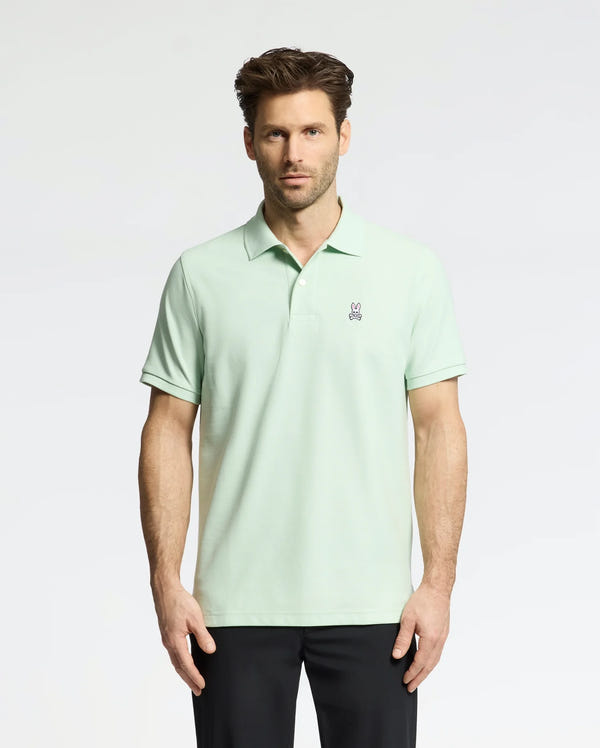 A man wears a Psycho Bunny Men's Classic Pique Polo (B6K001F200) in light green with an embroidered bunny logo on the chest, paired with black pants. He has short, styled hair and a neutral expression against a plain, light-colored background.