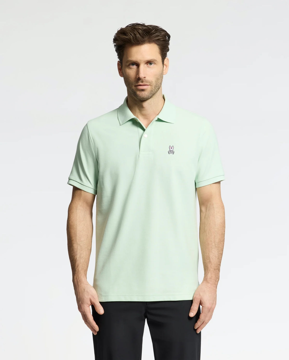 A man wears a Psycho Bunny Men's Classic Pique Polo (B6K001F200) in light green with an embroidered bunny logo on the chest, paired with black pants. He has short, styled hair and a neutral expression against a plain, light-colored background.