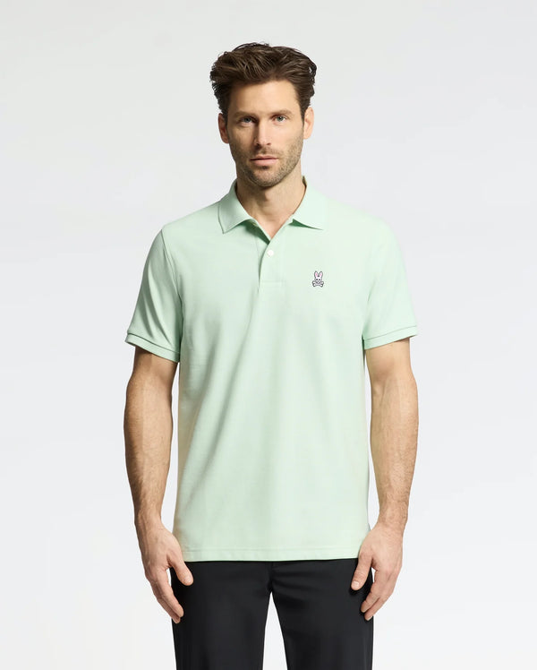 A man wears a Psycho Bunny Men's Classic Pique Polo (B6K001F200) in light green with an embroidered bunny logo on the chest, paired with black pants. He has short, styled hair and a neutral expression against a plain, light-colored background.