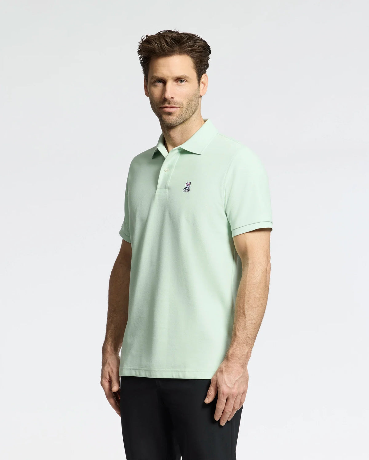 A man stands against a light background wearing the Psycho Bunny Men's Classic Pique Polo (B6K001F200) in light green with an embroidered bunny logo on the chest and black pants. He has short brown hair, facial hair, and looks confidently at the camera.