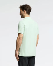 A dark-haired man stands with his back to the camera, wearing a light green Psycho Bunny Men's Classic Pique Polo (B6K001F200) with an embroidered bunny logo and black pants. The background is plain white.