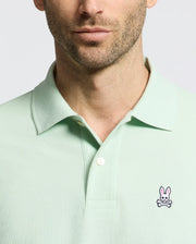 A man wears a light green Psycho Bunny Men's Classic Pique Polo (B6K001F200) featuring a pink and white embroidered bunny logo on the left chest, set against a plain neutral background that accentuates the polo's refined details.