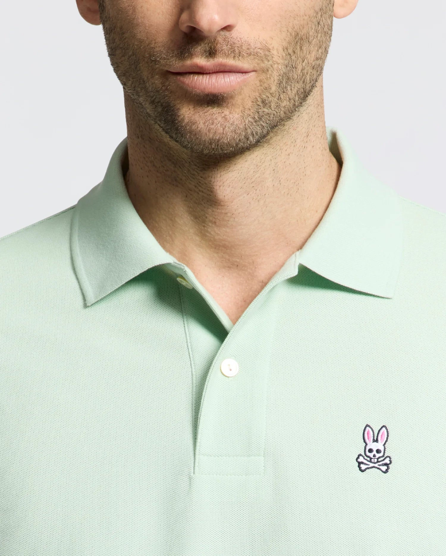 A man wears a light green Psycho Bunny Men's Classic Pique Polo (B6K001F200) featuring a pink and white embroidered bunny logo on the left chest, set against a plain neutral background that accentuates the polo's refined details.