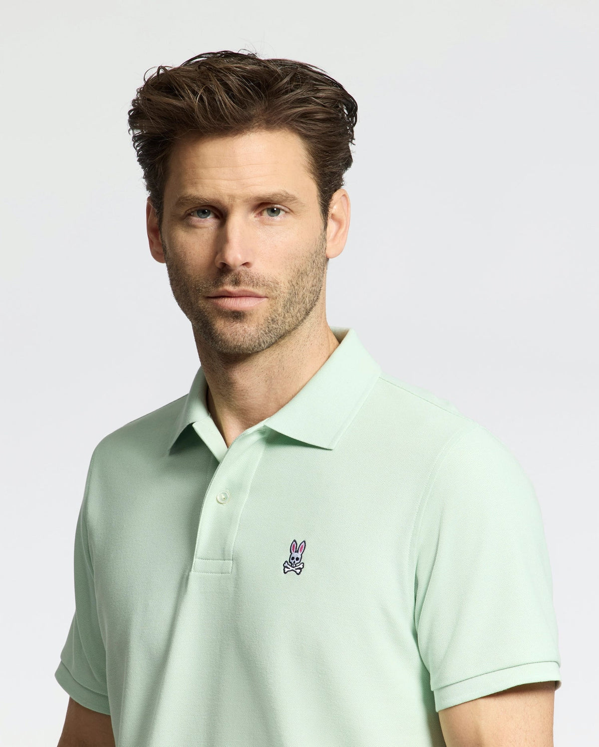 A brown-haired, bearded man wears the Psycho Bunny MENS CLASSIC PIQUE POLO - B6K001F200 in light green Pima cotton, with a small embroidered bunny logo on the chest. The backdrop is plain and light-colored.