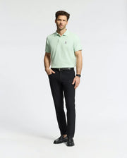 A person confidently wears a light green Psycho Bunny Men's Classic Pique Polo (B6K001F200) crafted from Pima Cotton with an embroidered bunny logo. Paired with black trousers and shoes, their hands rest casually in their pockets, short hair completing the look against a plain white background.
