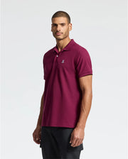 A man stands against a plain background, wearing a maroon Psycho Bunny Men's Classic Pique Polo Shirt (B6K001Z1PC), featuring an embroidered Bunny logo on the chest, and black pants. He has short hair and a beard, looking confidently at the camera.