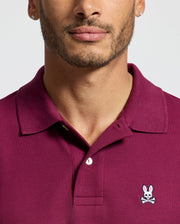 Someone is wearing a Psycho Bunny MENS CLASSIC PIQUE POLO SHIRT - B6K001Z1PC in burgundy, made from smooth Pima cotton with an embroidered Bunny logo on the chest, against a light gray background. The face is only partially visible.