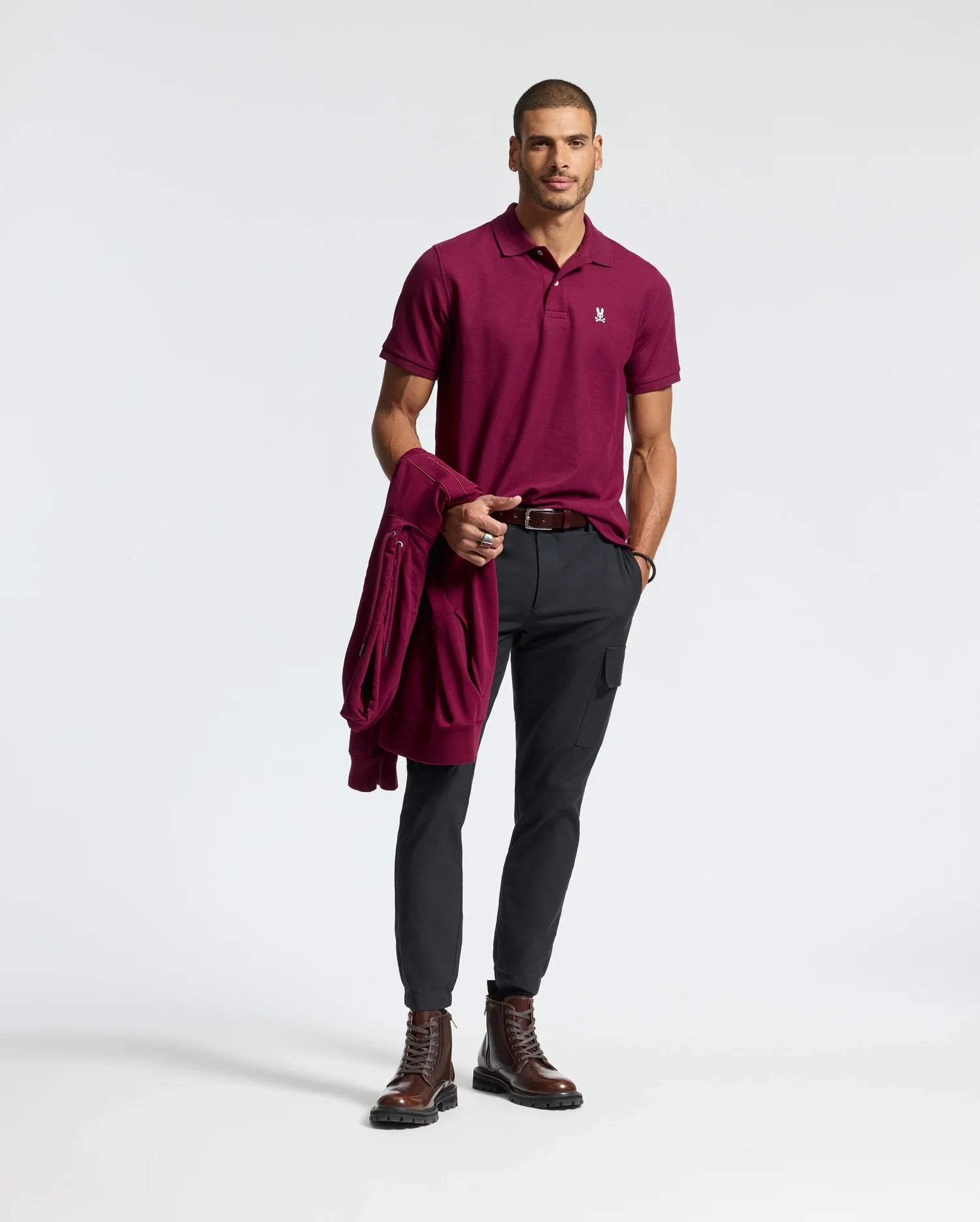 A man wearing a Psycho Bunny MENS CLASSIC PIQUE POLO SHIRT in burgundy, paired with black pants, stands holding a matching jacket. He completes his look with brown boots and a belt featuring an embroidered Bunny logo, set against a plain white background.