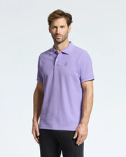 A man wearing a lavender Psycho Bunny men's Classic pique polo (B6K001Z1PC) with an embroidered Bunny logo on the chest stands against a plain white background. He has short, styled hair and a trimmed beard.
