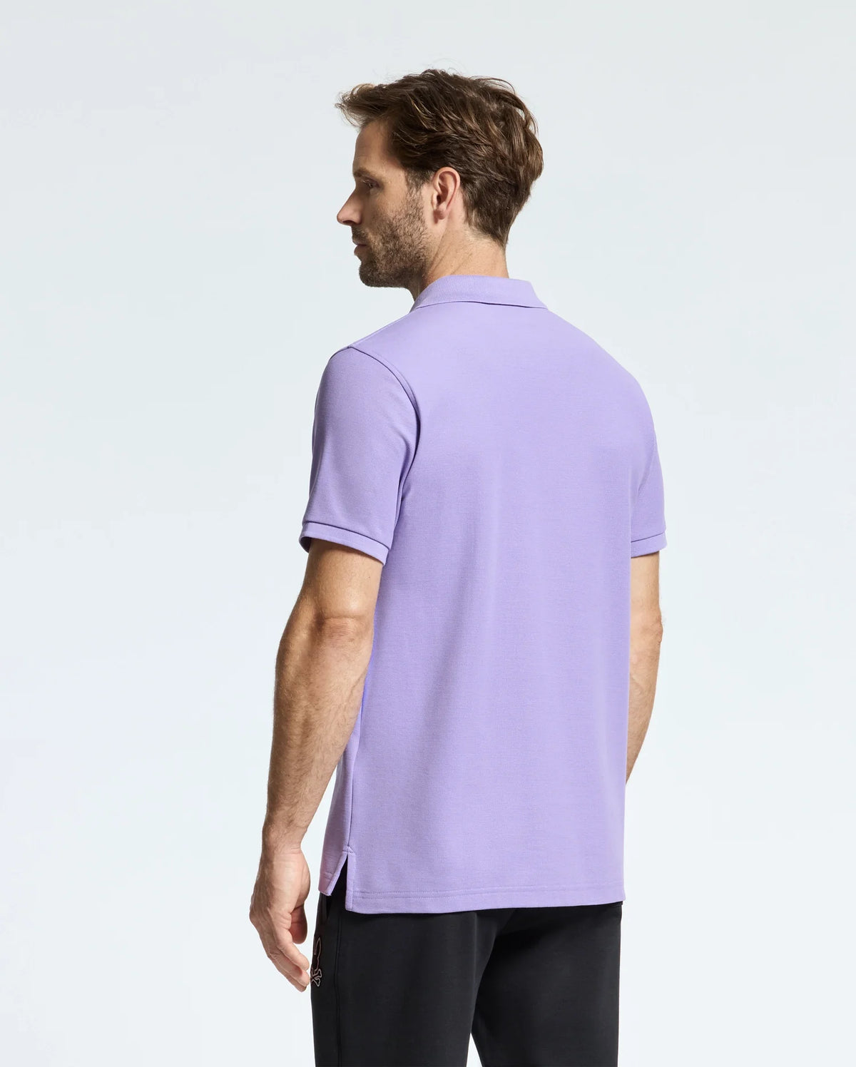 A person with short hair wearing a light purple Psycho Bunny men's Classic pique polo (B6K001Z1PC), featuring an embroidered Bunny logo, and dark pants is standing and facing away from the camera against a plain background.