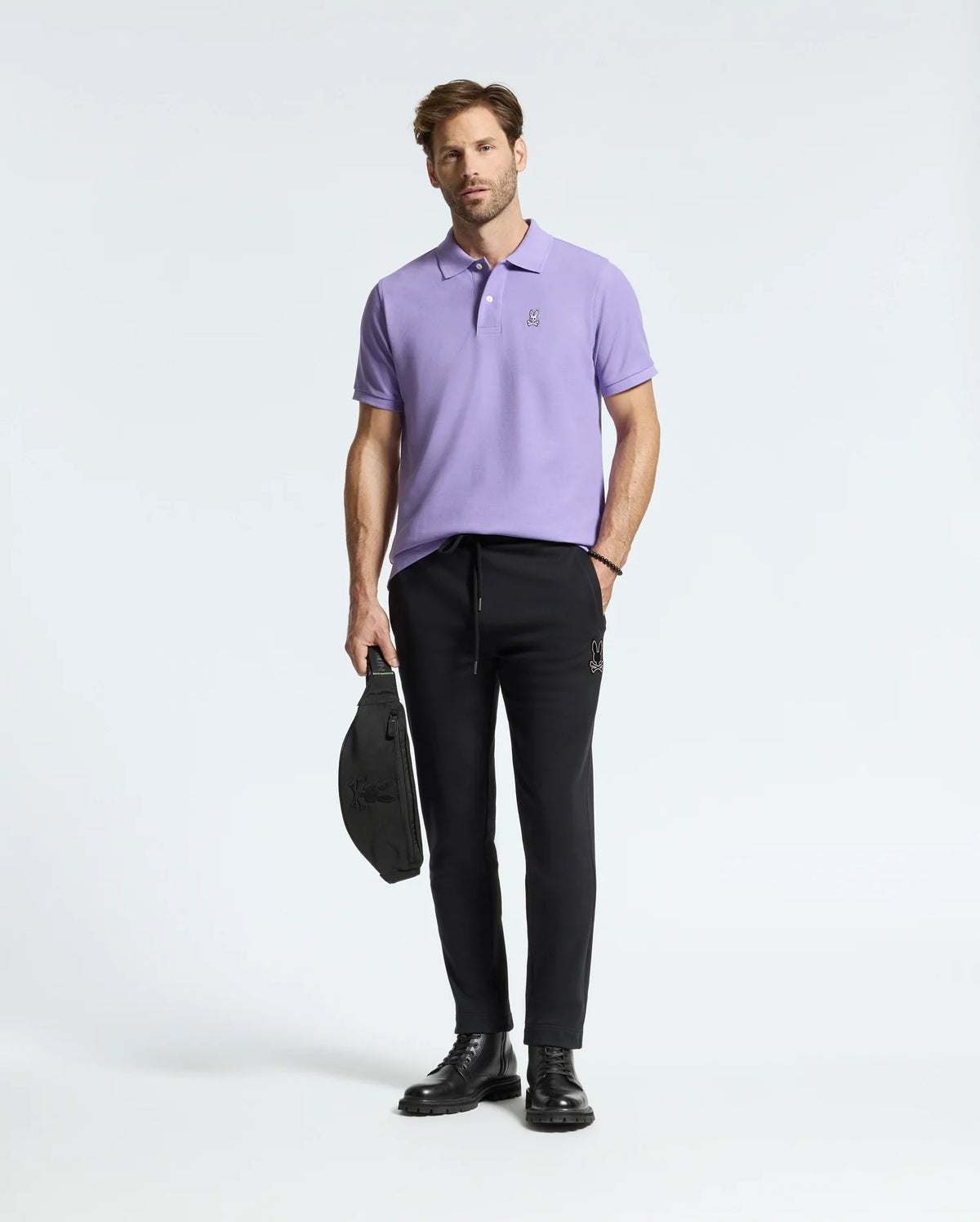A man wearing a Psycho Bunny men's Classic Pique Polo (product code: B6K001Z1PC) in lavender, paired with black jogger pants and black boots, is holding a black bag. He poses against a plain white background.