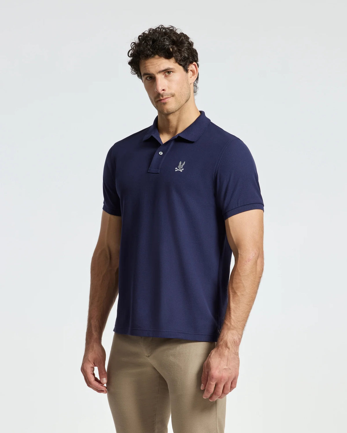 A man with curly hair stands against a light grey background wearing the Psycho Bunny MENS ALEXANDER PIQUE POLO - B6K101E200 in dark blue and a pair of beige pants. The polo, crafted from soft Pima cotton, showcases a small embroidered logo on the chest.