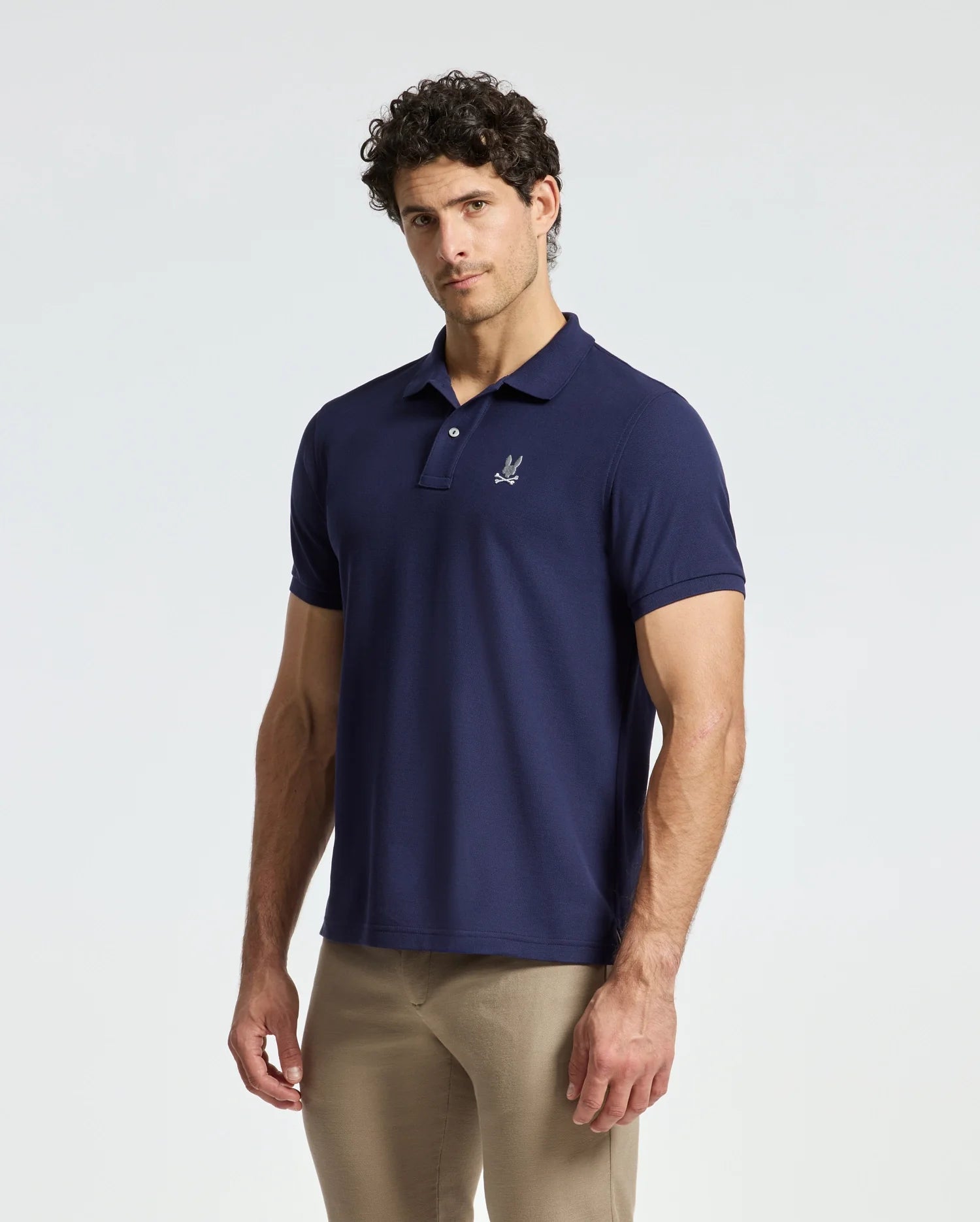 A man with curly hair stands against a light grey background wearing the Psycho Bunny MENS ALEXANDER PIQUE POLO - B6K101E200 in dark blue and a pair of beige pants. The polo, crafted from soft Pima cotton, showcases a small embroidered logo on the chest.