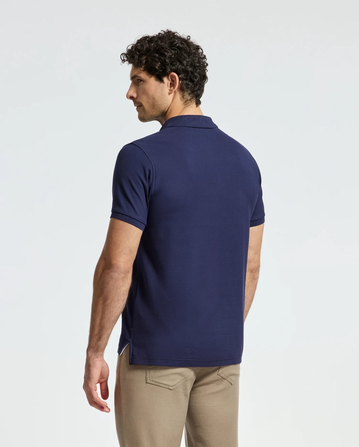 A man in a navy blue MENS ALEXANDER PIQUE POLO by Psycho Bunny stands with his back to the camera. He has short, curly hair and wears beige pants, slightly turned to the side against a plain, light background. The classic Alexander style exudes understated elegance and comfort.
