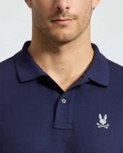 A person wears a dark blue Men's Alexander Pique Polo from Psycho Bunny, showcasing a small embroidered rabbit's head and crossbones logo on the chest. The image centers on the upper torso and neck against a plain background, emphasizing this stylish pique polo design.