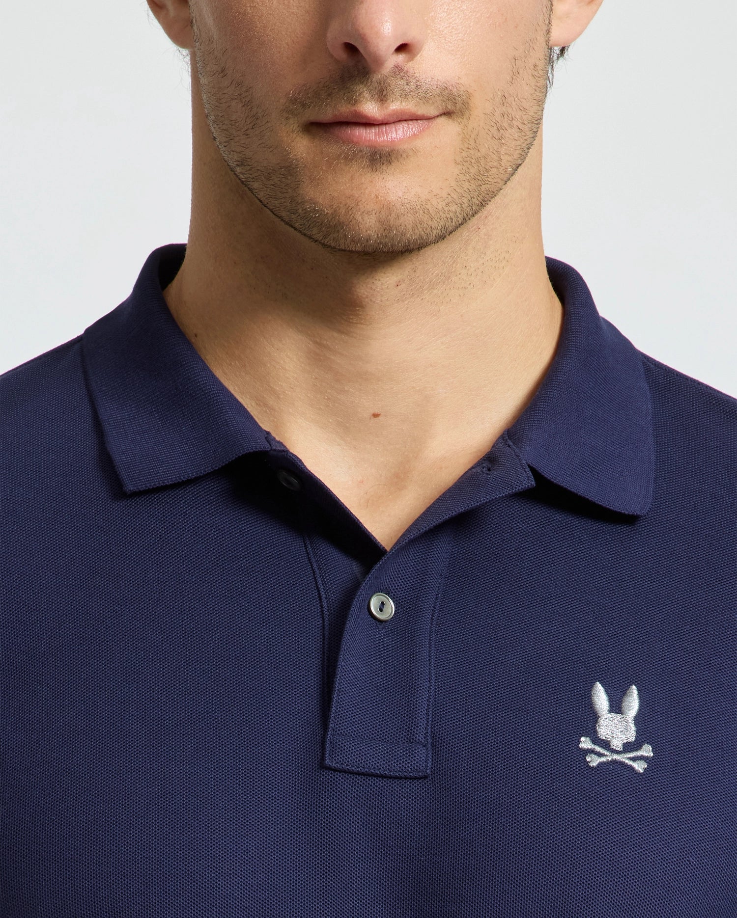 A person wears a dark blue Men's Alexander Pique Polo from Psycho Bunny, showcasing a small embroidered rabbit's head and crossbones logo on the chest. The image centers on the upper torso and neck against a plain background, emphasizing this stylish pique polo design.