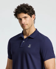 A man with curly hair is wearing a dark blue MENS ALEXANDER PIQUE POLO by Psycho Bunny, which showcases a small embroidered eagle emblem on the left side of the chest. The background is a plain, light color.